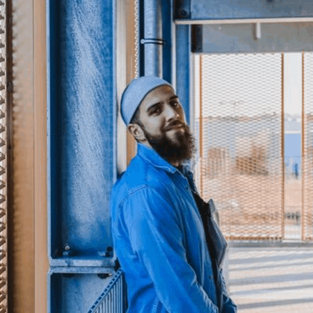 a man with a beard wearing a hat and a blue jacket is leaning against a blue wall .