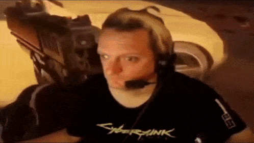 a man wearing headphones and a cyberpunk 2077 t-shirt is sitting in front of a car .