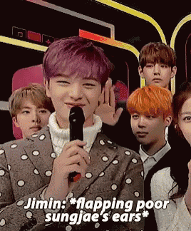 a man with purple hair is holding a microphone in front of a group of people including sungjae 's ears .