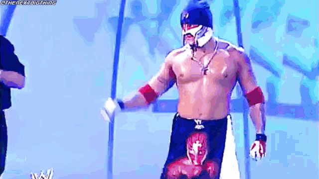 a shirtless wrestler wearing a mask and gloves is standing in front of a blue wall .