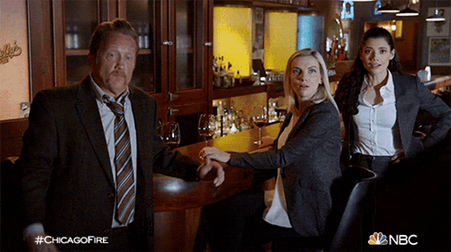 a man in a suit and tie stands next to a woman sitting at a bar with a nbc logo on the bottom