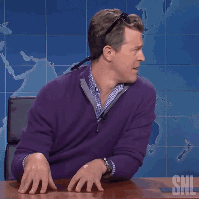 a man in a purple sweater is sitting at a table with a snl logo on the desk