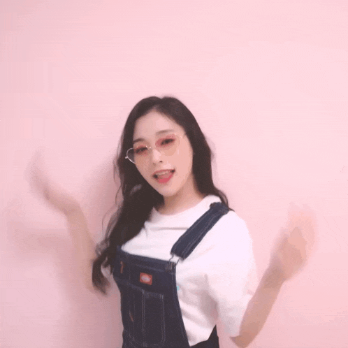a woman wearing overalls and a white t-shirt is standing in front of a pink wall .