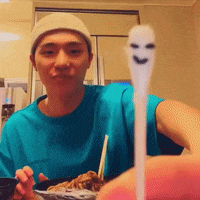 a man wearing a beanie and a blue shirt is eating a meal with a spoon with a face on it