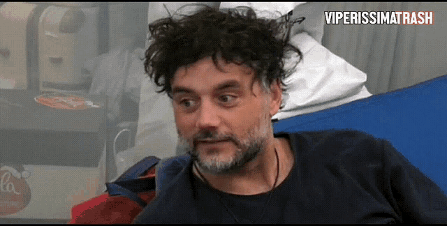 a man with a beard and curly hair is sitting on a bed with the words viperissima trash written on the bottom