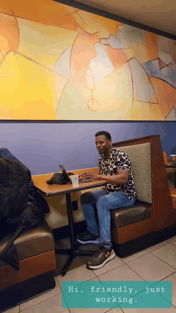 a man sits at a table with a laptop and a cup of coffee and says " hi friendly just working "