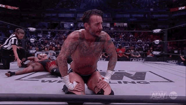a wrestler is kneeling down in a wrestling ring while another wrestler lays on the ground .