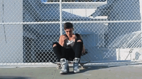 a young man sits on a skateboard in front of a chain link fence smoking a cigarette