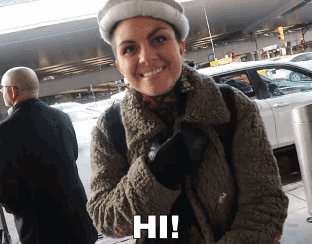 a woman wearing a hat and a jacket is smiling and says hi