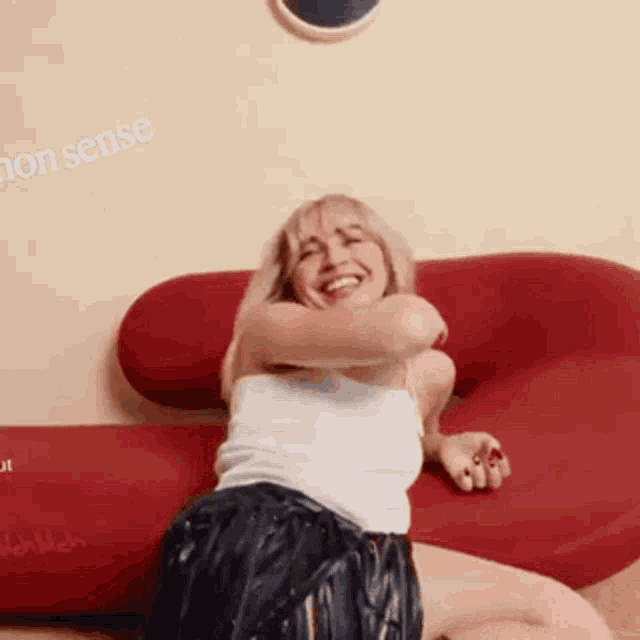 a woman in a white tank top and black shorts is sitting on a red couch and smiling .