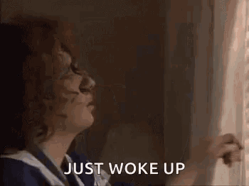 a woman is looking out of a window while holding a blind and says `` just woke up '' .