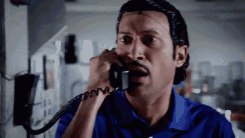a man in a blue shirt is talking on a phone with a surprised look on his face