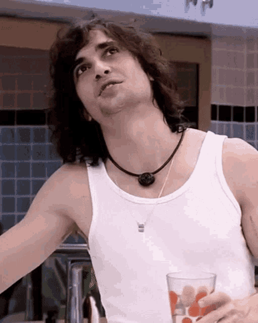 a man with long hair is wearing a white tank top and holding a glass