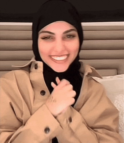 a woman wearing a hijab and a trench coat is smiling with her hands folded .