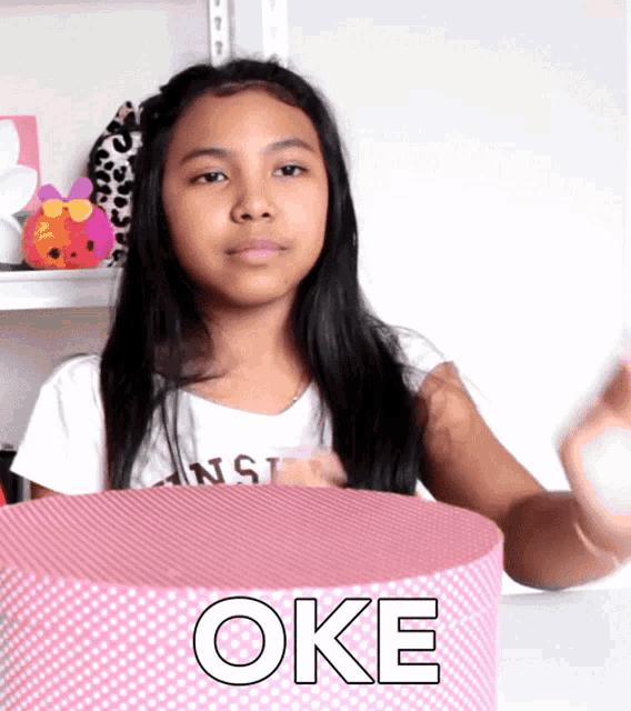 a young girl is sitting in front of a pink box that says oke