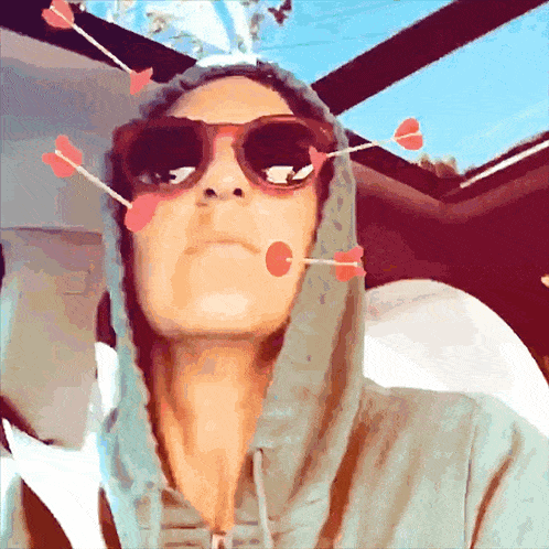 a woman wearing sunglasses and a hoodie has arrows stuck to her face