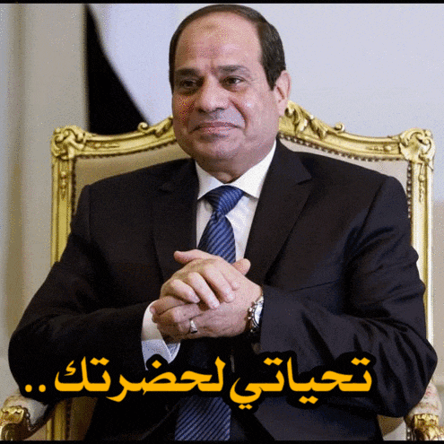 a man in a suit and tie is sitting in a chair with arabic writing behind him