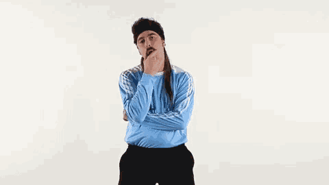 a man wearing a mullet and a headband is standing with his arms crossed and his hand on his chin .