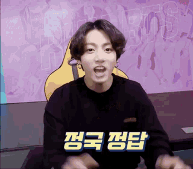 a man in a black sweater is making a funny face in front of a purple wall with korean writing .