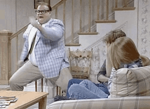a man in a plaid jacket is dancing in front of a group of people sitting on a couch in a living room .