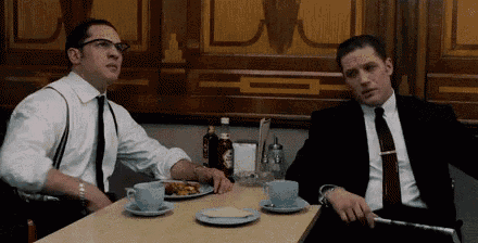 two men in suits and ties are sitting at a table with cups of coffee .