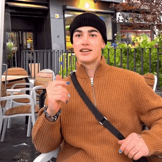 a man wearing a beanie and a brown sweater is sitting outside