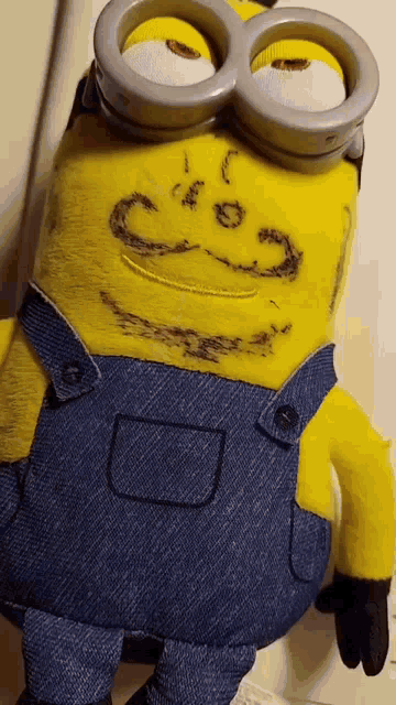 a stuffed minion wearing overalls and goggles has a smiley face drawn on it