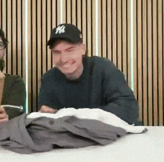 a man wearing a ny hat sits at a table with a pile of clothes