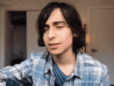 a young man with long black hair is wearing a plaid shirt and a blue shirt .
