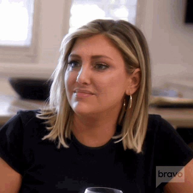 a woman is sitting at a table with a glass of wine and a bravo logo on the bottom of her shirt