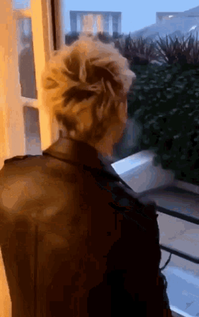 a man in a black leather jacket is standing in front of a window looking out .