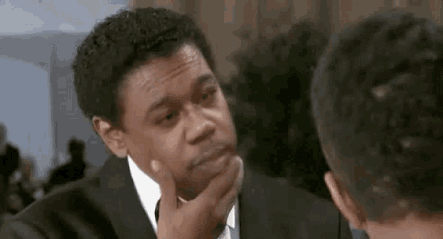 a man in a suit is talking to another man while holding his hand to his chin .