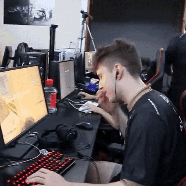 a man wearing a black shirt with the word assault on the back sits at a desk playing a video game