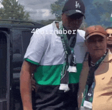 a man in a la hat stands next to another man in a green and white shirt