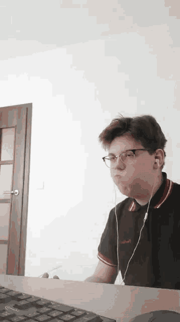 a young man wearing ear buds and a black shirt with red and white stripes is sitting at a desk with a keyboard