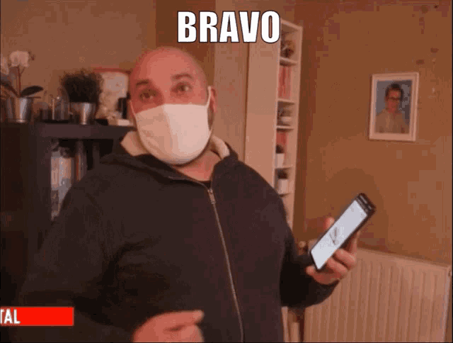 a man wearing a face mask holds a cell phone with the word bravo above him