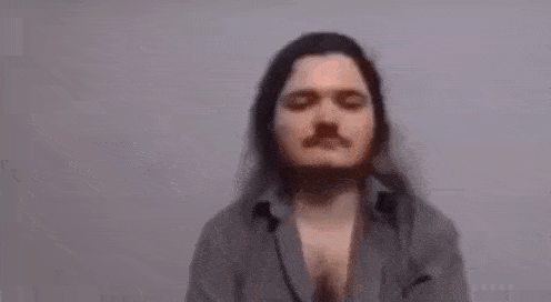 a man with long hair and a beard is sitting in front of a white wall with his eyes closed .
