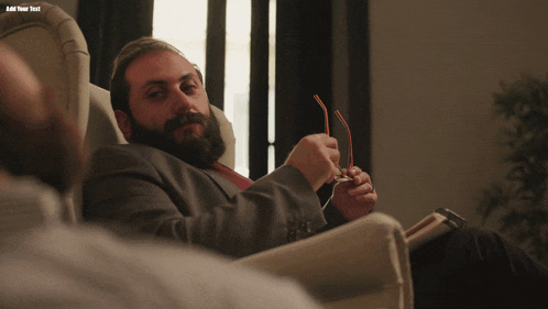 a man with a beard sits in a chair holding his glasses