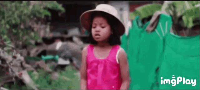 a little girl wearing a pink dress and a hat is standing in front of a green fence .