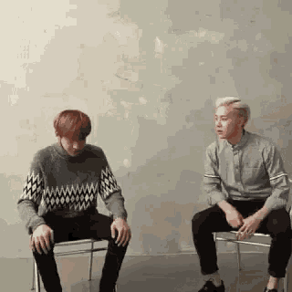 two men are sitting in chairs in front of a wall talking to each other .