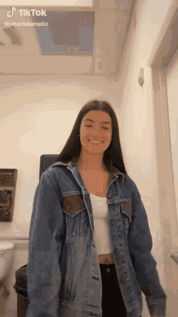 a woman wearing a denim jacket and a white tank top is smiling in a room .