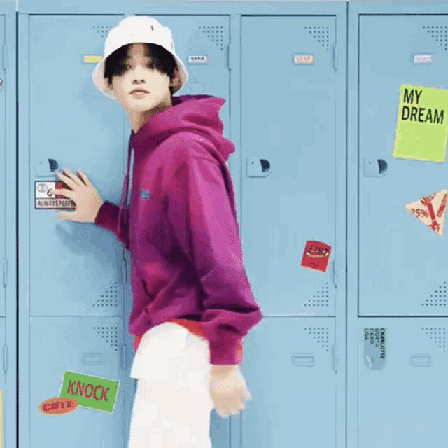 a young man wearing a pink hoodie and a white hat is standing in front of a row of blue lockers .