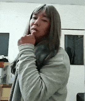 a woman with long hair is standing in a room with her hand on her chin and looking at the camera .