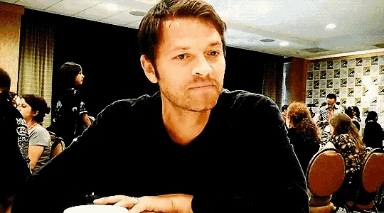 a man in a black shirt is sitting at a table in a room with a group of people .