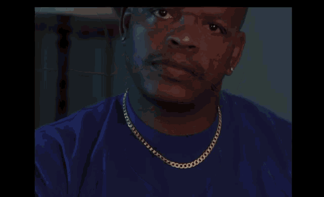 a bald man wearing a blue shirt and gold chains looks at the camera