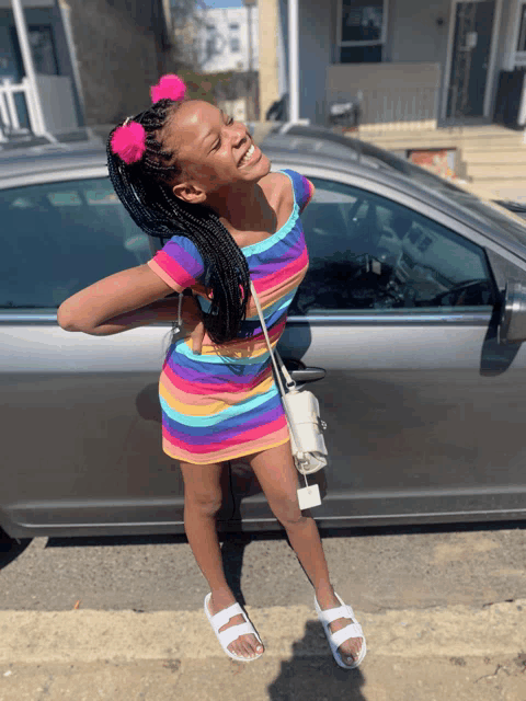 a young girl wearing a rainbow dress and white sandals stands in front of a car