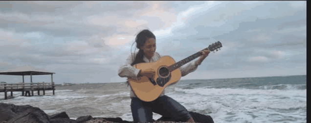 a woman is kneeling on a rock playing a guitar by the ocean