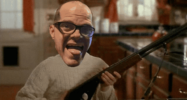 a bald man wearing glasses is holding a shotgun and sticking his tongue out