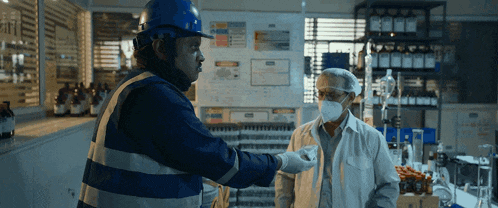 a man wearing a blue hard hat talks to another man wearing a mask