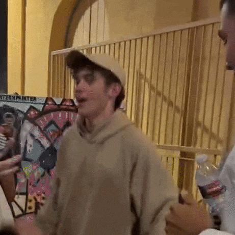 a man wearing a baseball cap and a hoodie is talking to another man while holding a bottle of water .